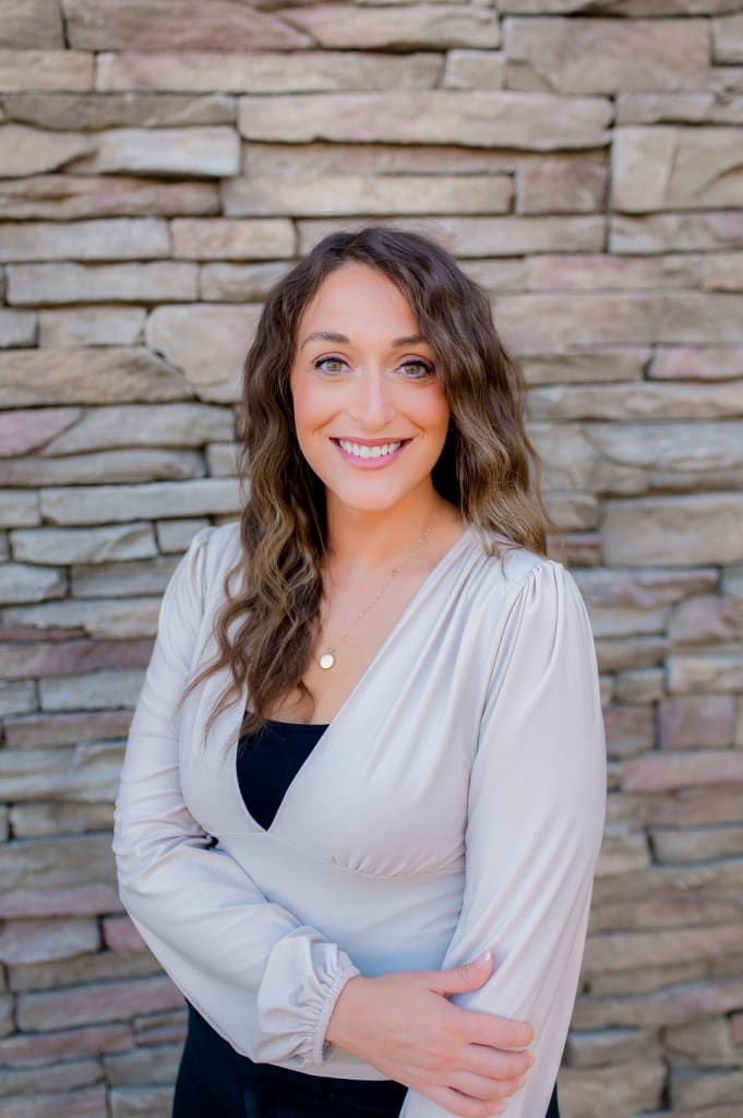 A woman with long wavy hair and a light blouse smiles while standing in front of a stone wall.